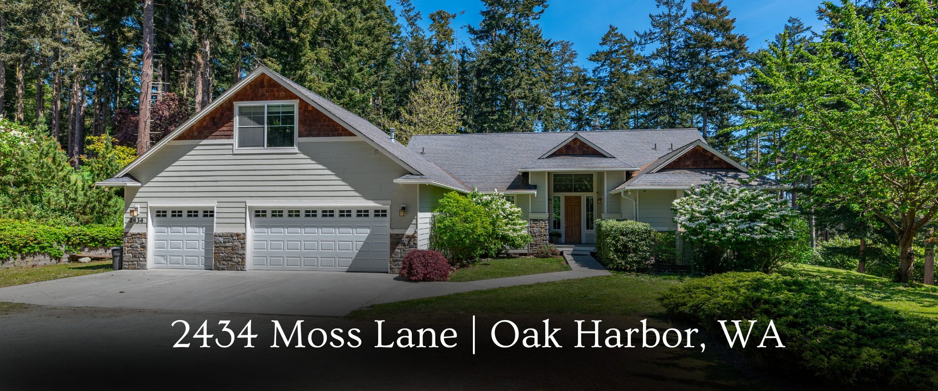 Exterior view of 2434 Moss Lane, a contemporary contemporary one-story home in Oak Harbor, WA. The residence features a three-car garage, stone accents, and a wide driveway, nestled on a private, wooded one-acre lot near Joseph Whidbey State Park.